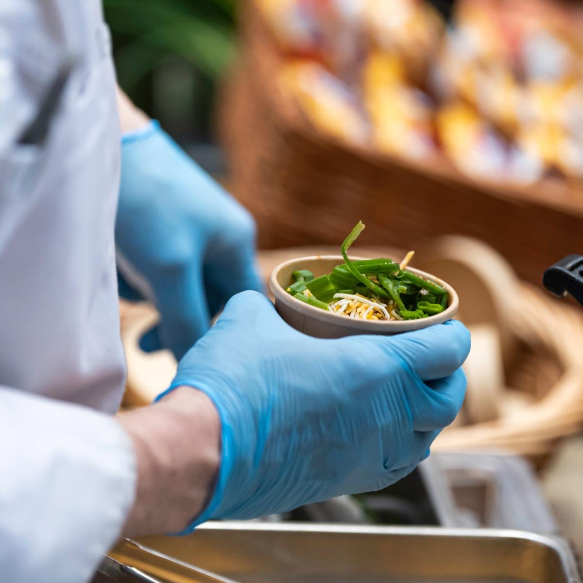 Gloved hands serve a cup of chili garnished with green onions and cheese on top